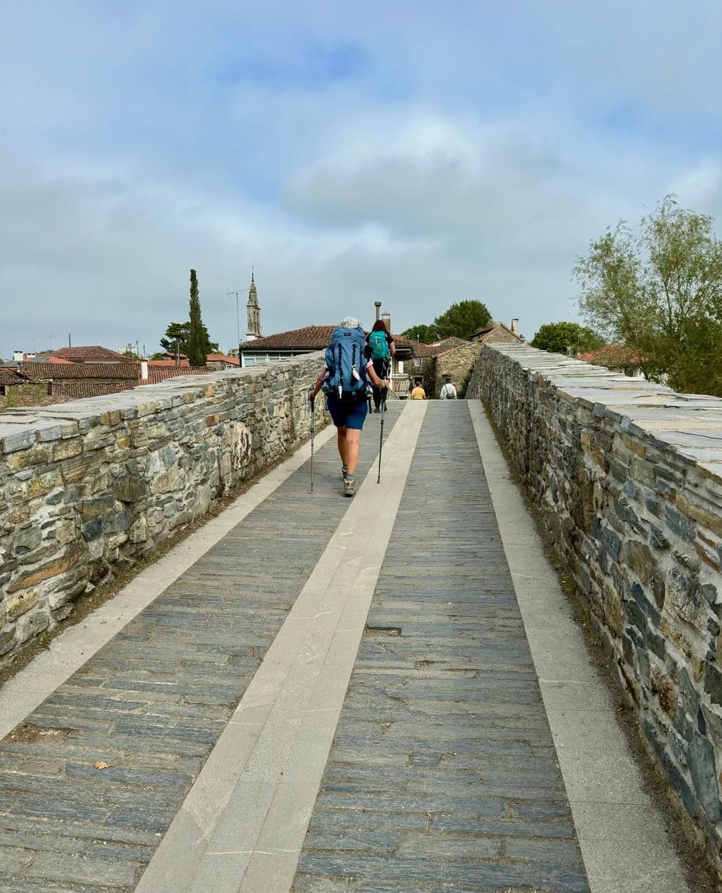 Deux pèlerins sac au dos et bâtons de marche traversant un pont de pierre médiéval à l'entrée d'un village galicien