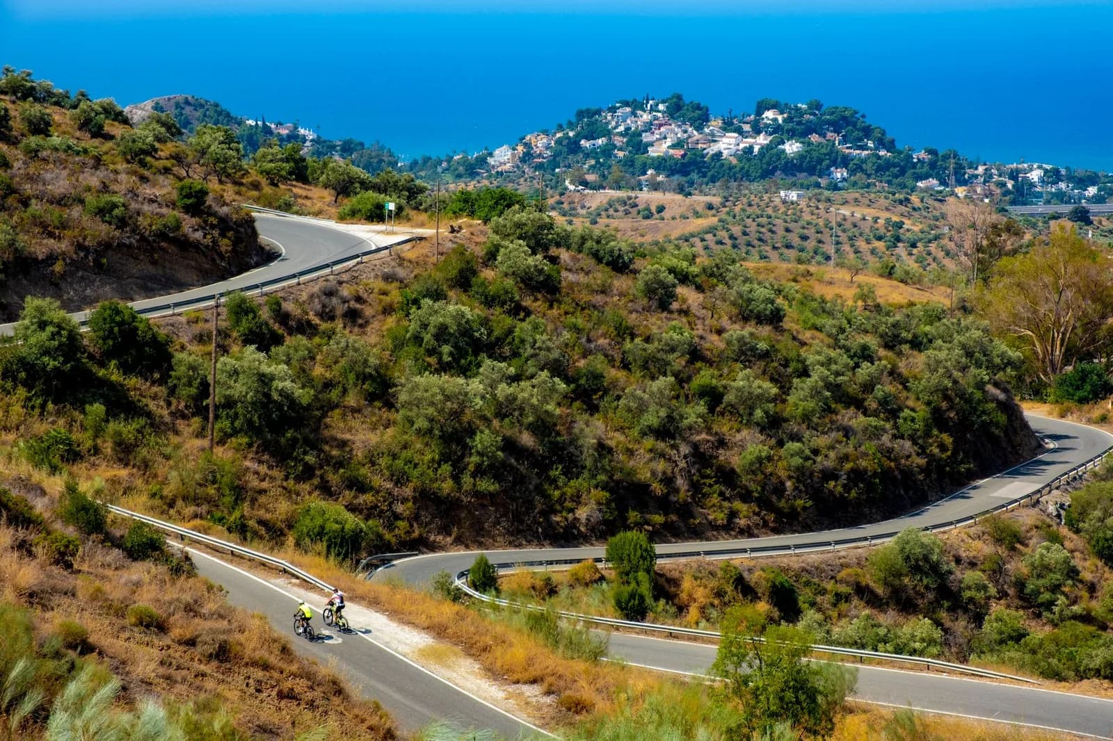 Route sinueuse de la côte andalouse au-dessus de la Méditerranée, oliveraies et village blanc sur la colline, deux cyclistes au premier plan