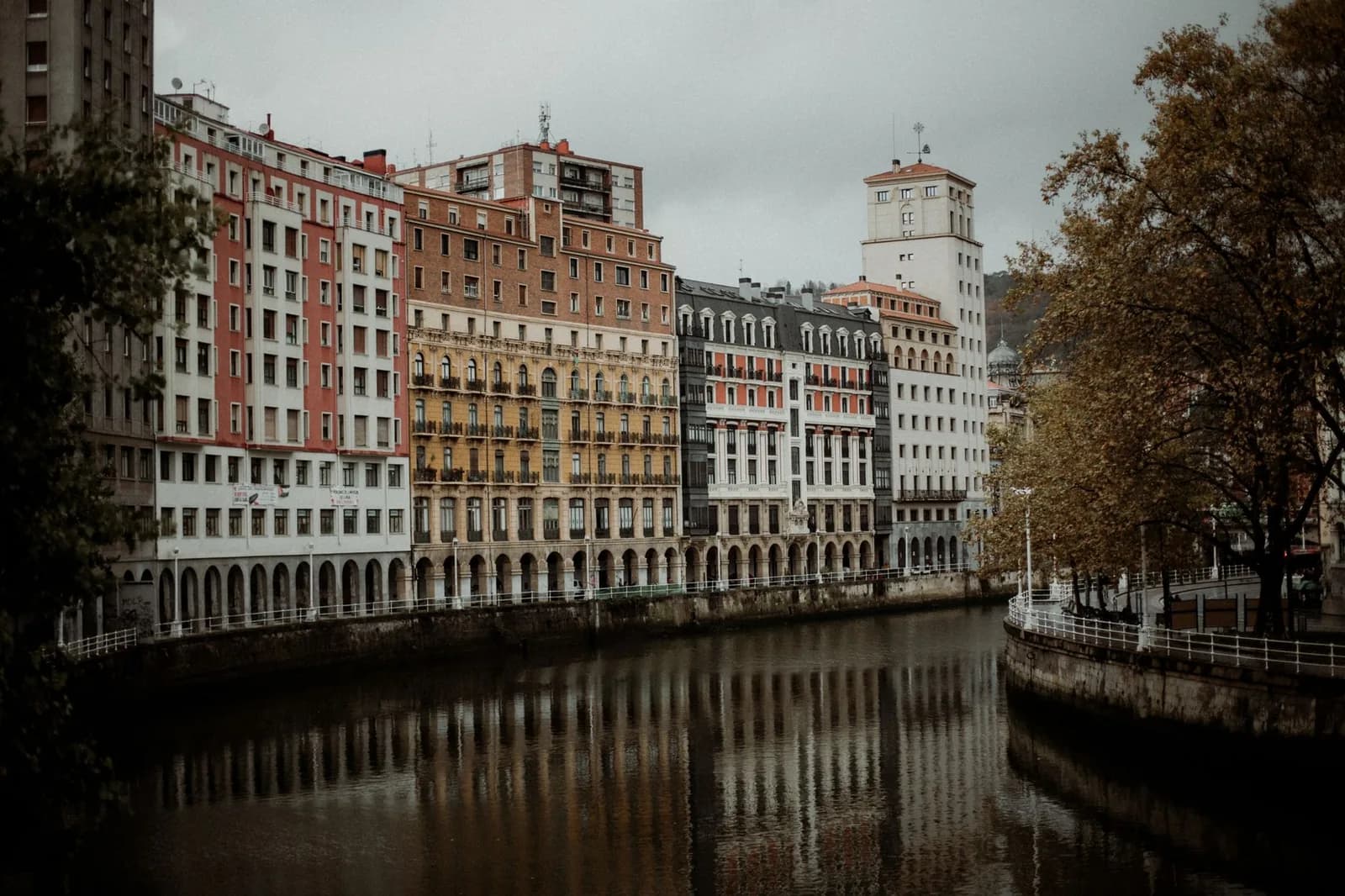 Les quais arcadés du Nervión à Bilbao sous un ciel gris d'automne, immeubles colorés reflétés dans l'eau