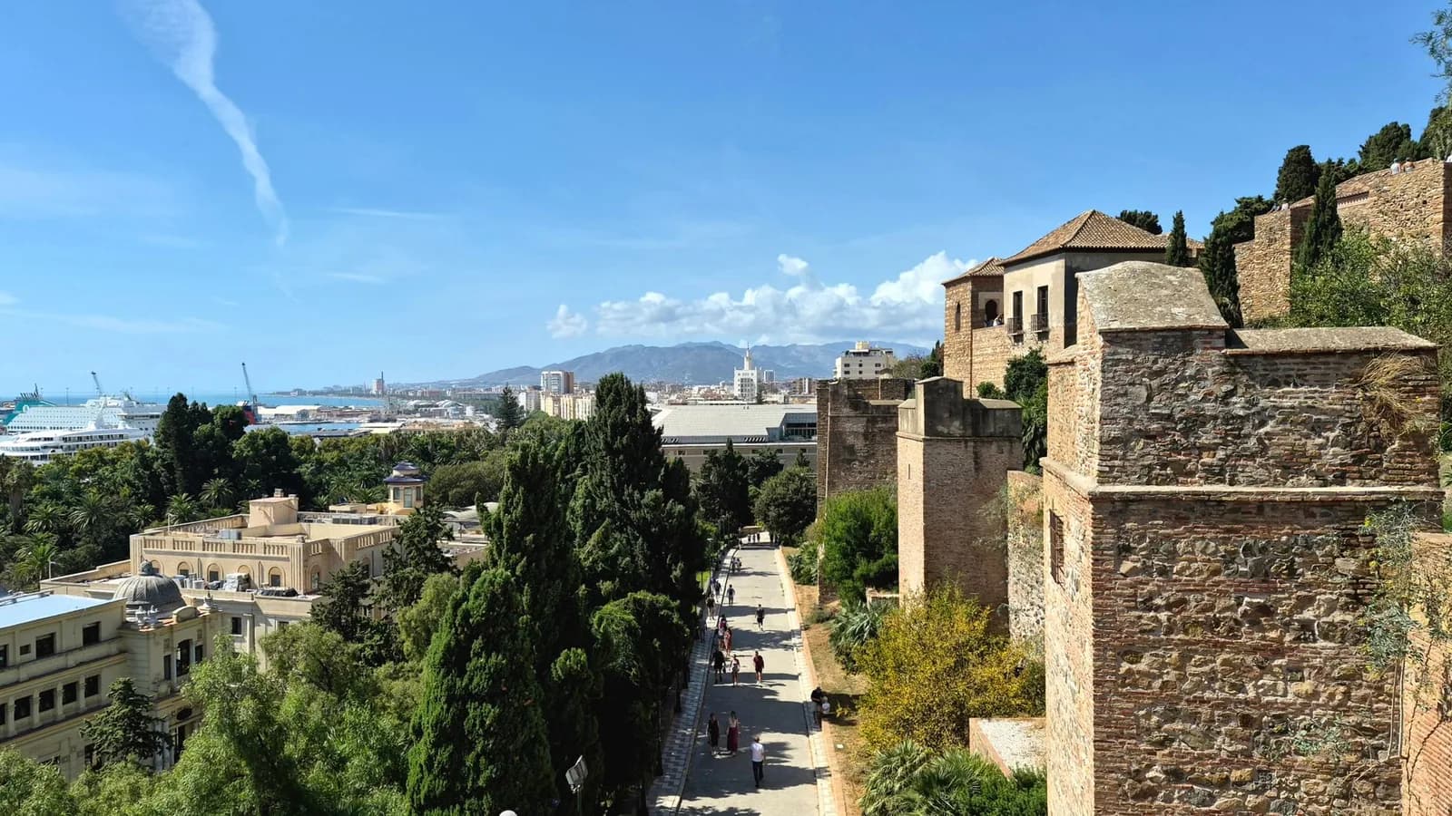 Remparts et tours de l'Alcazaba de Málaga dominant une allée bordée de cyprès, avec le port et les montagnes en arrière-plan