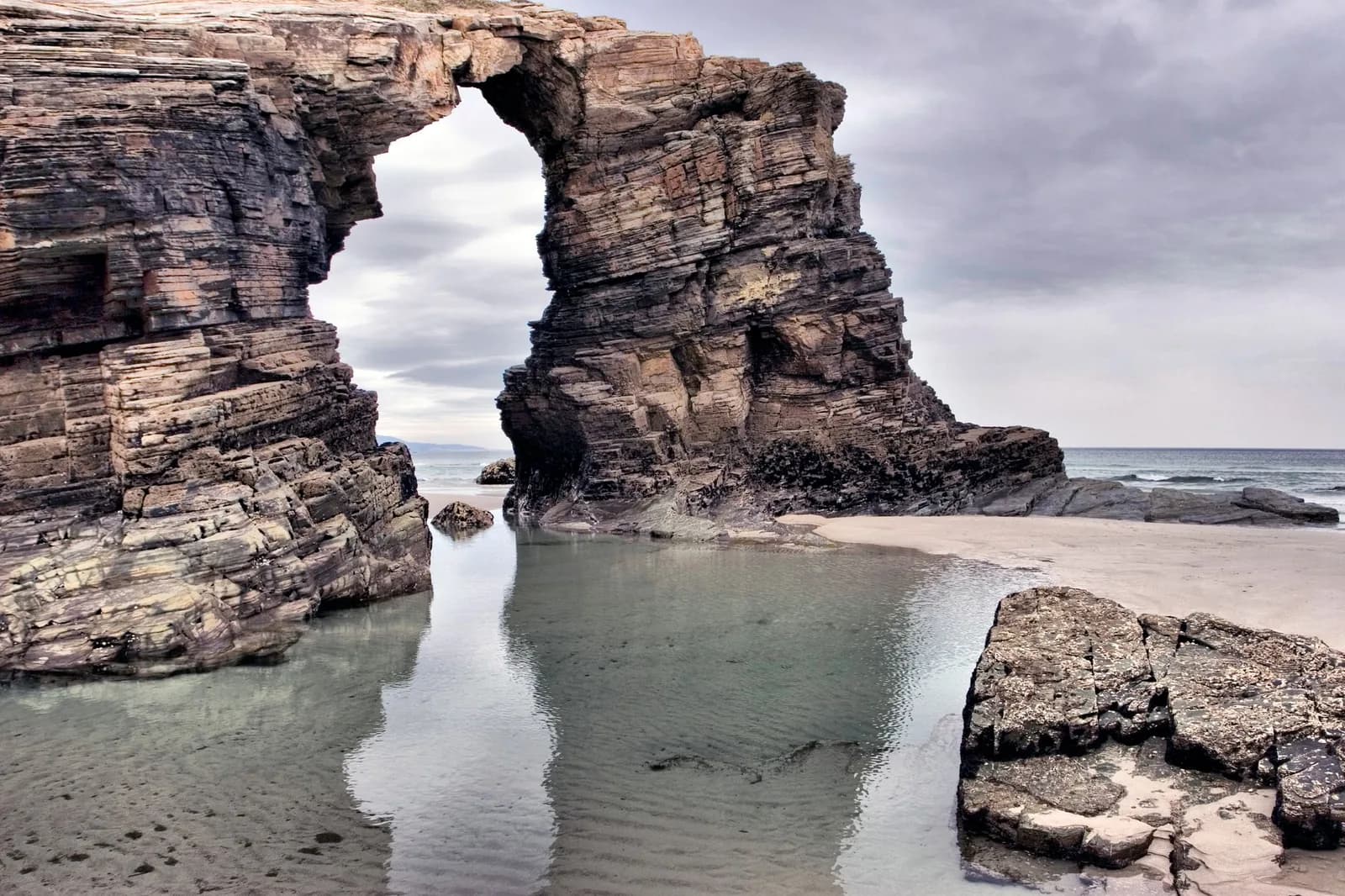 L'arche de schiste de la Playa de las Catedrales en Galice, à marée basse, devant une vasque d'eau et le sable